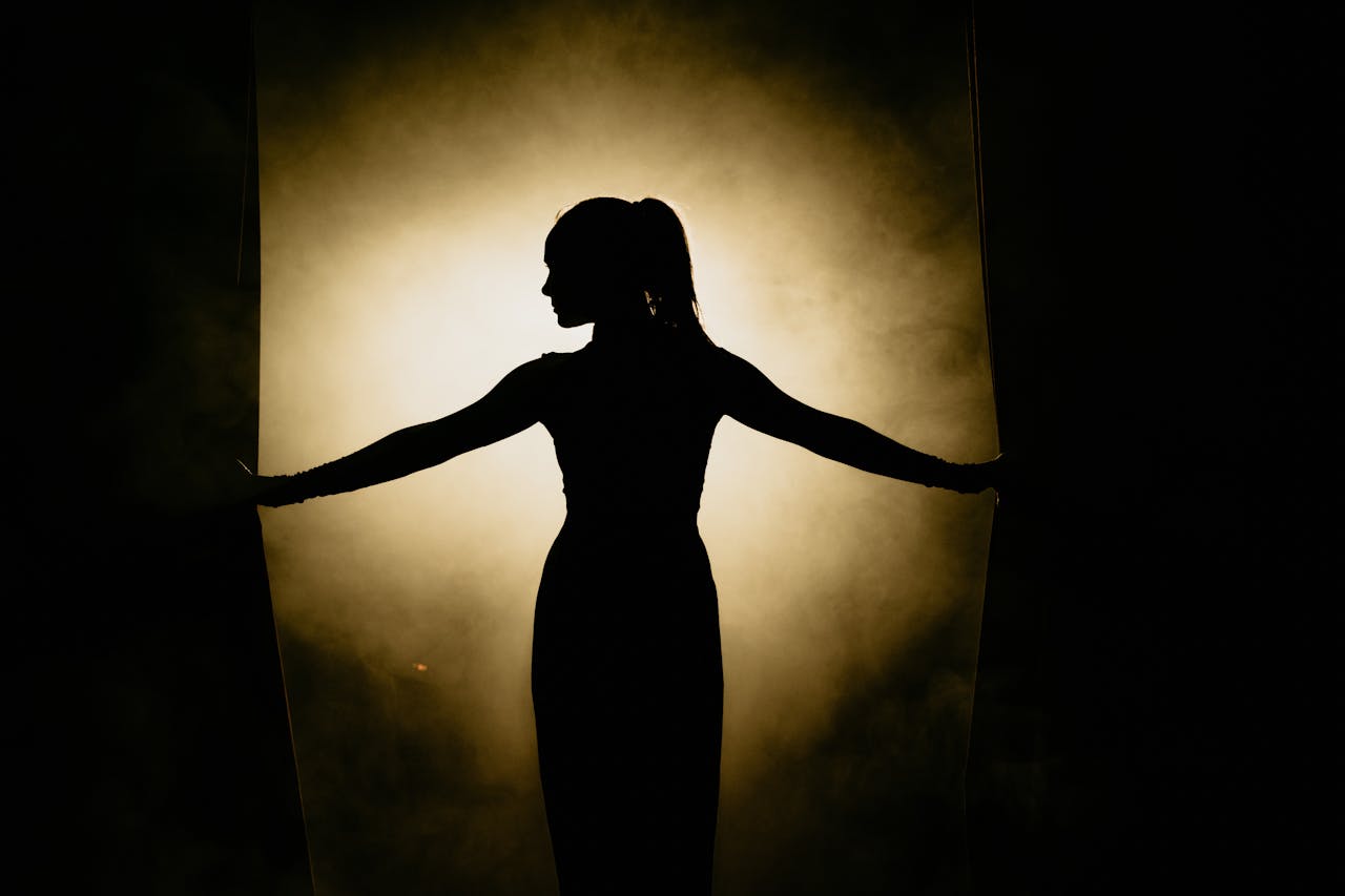 about-img Artistic silhouette of a woman opening stage curtains, surrounded by smoke and spotlight.